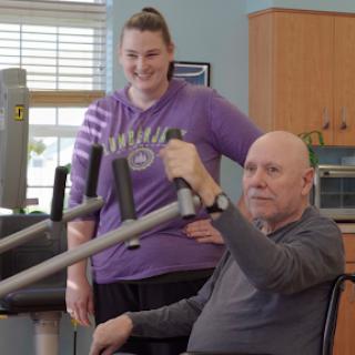 A senior man engages in a strength training exercise with the assistance of a trainer in a rehabilitation setting.
