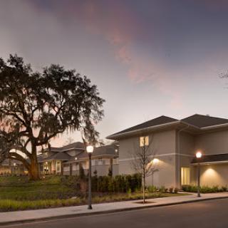 A modern residential building illuminated at dusk, surrounded by landscaping and an oak tree.