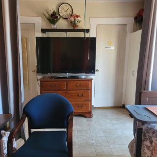 A cozy living room with a television on top of a wooden dresser, a navy blue chair in the foreground, and decorative elements including a clock and flowers.