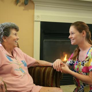 A caregiver shares a warm moment with an elderly woman by the fireplace.
