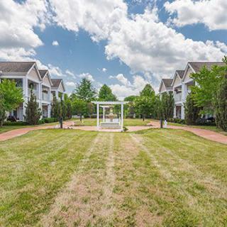 A serene landscape featuring a central gazebo surrounded by grassy lawns and buildings under a partly cloudy sky.