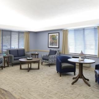 A comfortably furnished living room with sofas, armchairs, and tables arranged neatly, featuring large windows and a cozy carpet.