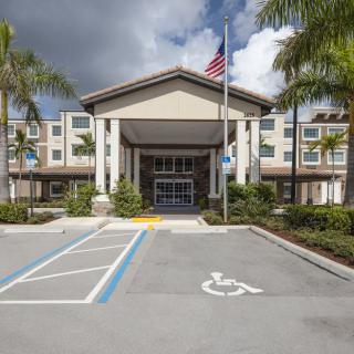 Entrance of a building with palm trees and an American flag, featuring accessibility parking in front.