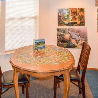 A cozy room with a wooden table featuring an incomplete jigsaw puzzle, surrounded by chairs and decorated with artwork on the walls.