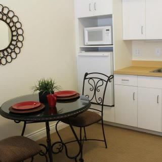 A compact kitchen and dining area featuring a round table with two chairs, adorned with red plates. A microwave is set above white cabinets, complemented by a decorative mirror on the wall.