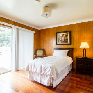 A cozy bedroom with wood paneling, featuring a single bed, nightstand with lamp, and sliding door leading outside.