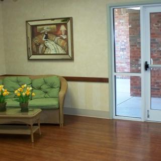 A cozy waiting room featuring a green sofa, a wooden coffee table with flowers, a painting on the wall, and a glass door leading outside.