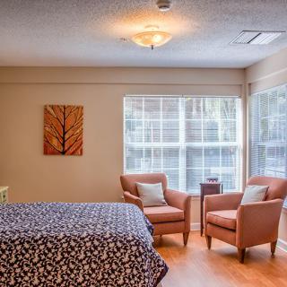 A cozy bedroom featuring a bed with patterned bedding, two armchairs by large windows, and a piece of artwork on the wall.