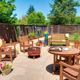 A sunny patio area with wooden furniture and a fire pit, surrounded by lush greenery.