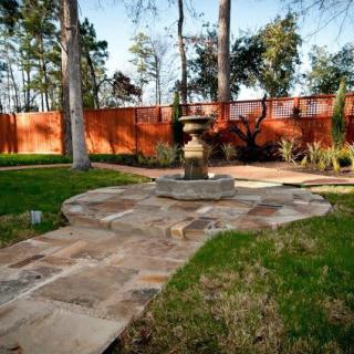 A tranquil garden scene with a stone path leading to a small fountain, surrounded by trees and enclosed by a wooden fence.