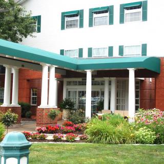 An exterior view of a building entrance with green canopies, columns, and landscaped garden.