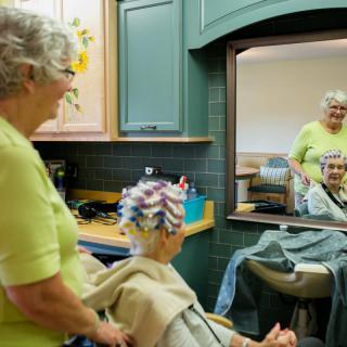 An elderly woman getting her hair styled in a salon, smiling at her reflection in the mirror.