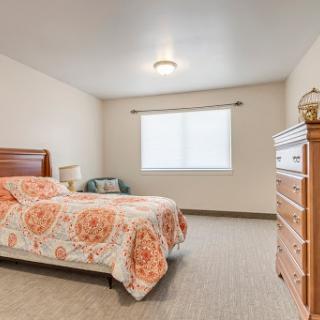 A cozy bedroom featuring a floral-patterned bedspread, wooden furniture, and natural light coming through the window.