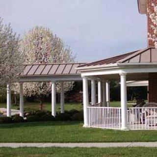 A covered walkway with white columns next to a brick building, surrounded by blooming trees.