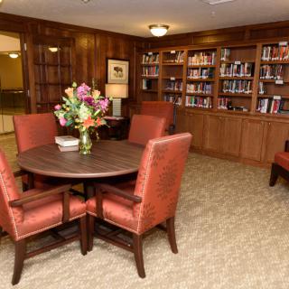 A cozy library with wooden bookshelves, a round table surrounded by upholstered chairs, and a vase of flowers on the table.