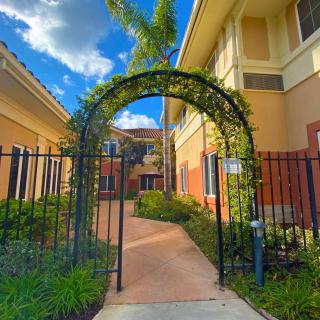 A scenic outdoor walkway with an archway covered in greenery, leading to a building under a bright blue sky.