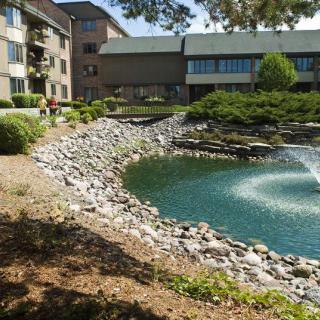 A tranquil scene with an apartment building overlooking a small lake with a fountain and rocky shoreline.