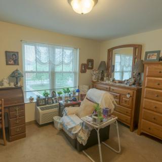 A cozy bedroom with a large window, featuring a comfortable chair, dresser, and various decorations.