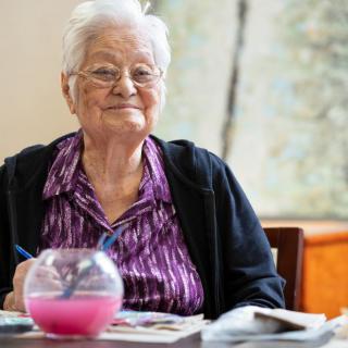 An elderly woman smiling while engaged in painting at a table.