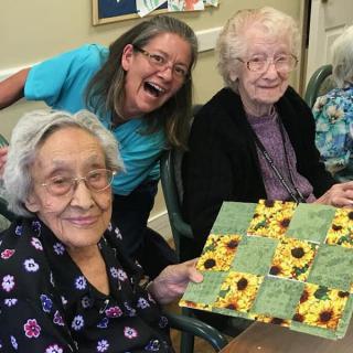 Three women smiling together, with one holding a sunflower-themed artwork.