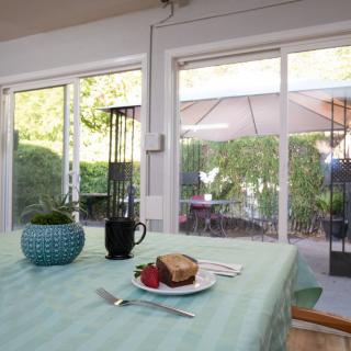 A table set with cake and a strawberry next to a coffee mug, overlooking a patio through large windows.