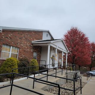 A brick building with an accessibility ramp, surrounded by autumn foliage under a cloudy sky.