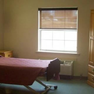 A simple room featuring a hospital bed, window with blinds, and wooden furniture.