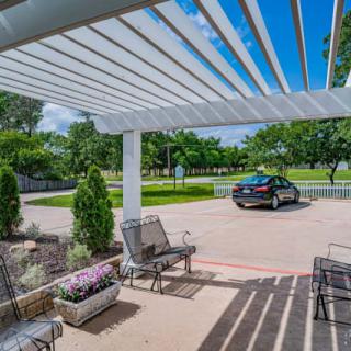 A sunny patio with a pergola, chairs, and a view of a parked car and greenery.
