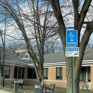 A blue handicap parking sign stands in front of a public building, surrounded by bare trees.