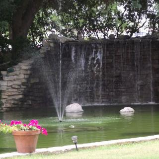 A tranquil garden scene with a fountain and waterfall by a pond, surrounded by trees and flowers.
