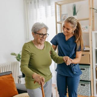 A nurse assists an elderly woman with a walking cane in a home setting.