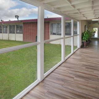 A covered walkway with glass walls and a wooden floor, leading to a brick building, adorned with indoor plants.