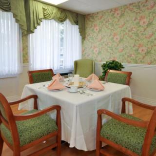 A neatly arranged dining table with green upholstered chairs in a room with floral wallpaper and curtains.