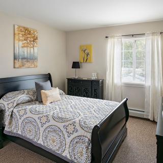 A cozy bedroom featuring a neatly made bed with patterned bedding, a dresser, and wall art. Natural light streams through the window with white curtains.