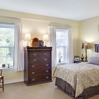 A cozy bedroom with traditional decor featuring a bed, chest of drawers, and two windows with curtains.