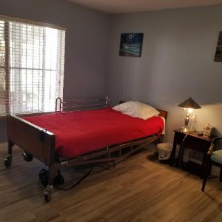A simple bedroom with a hospital bed featuring a red blanket, next to a nightstand with a lamp and window blinds partially open.