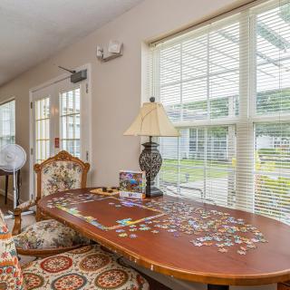 A cozy room with a wooden table covered in a partially completed puzzle, accompanied by an ornate lamp and patterned chair, bathed in natural light from large windows.