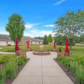A serene garden pathway leading to a fountain, surrounded by lush greenery and vibrant red umbrellas against a clear blue sky.