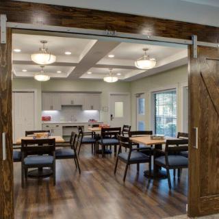 A modern dining area with wooden tables and chairs, connected to a kitchen space, featuring elegant lighting and wood flooring.