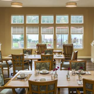 An empty dining room with neatly arranged tables and chairs, illuminated by natural light through large windows.