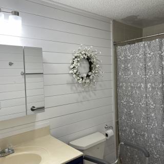 A well-lit bathroom featuring a mirror, decorative wreath on white wood paneling, and a patterned shower curtain.