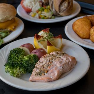 A meal featuring seasoned salmon, broccoli, and potatoes on a plate, with other dishes in the background.
