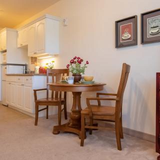 A cozy dining area with wooden furniture adjacent to a kitchen, featuring a small table with chairs and decorative elements like flowers and framed pictures on the wall.