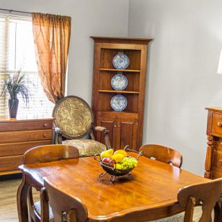 A cozy dining room with a wooden table and chairs, featuring a fruit bowl centerpiece. The corner cabinet displays decorative plates, and the window is adorned with sheer curtains.