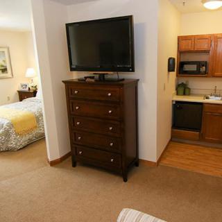A cozy hotel room featuring a bedroom with a television atop a dresser and a small kitchenette.