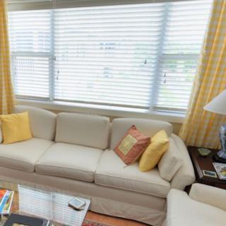 A cozy living room with a beige sofa adorned with colorful cushions, next to large windows with blinds and curtains.