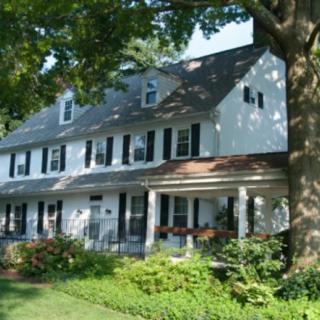 A large colonial-style house with a porch, surrounded by lush greenery and trees.
