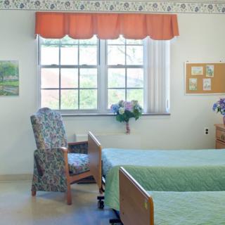 A well-lit room with two beds, a chair, and a window adorned with curtains.