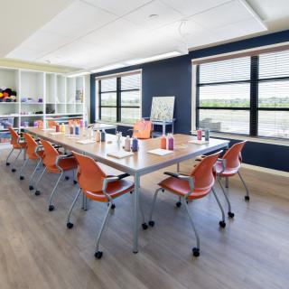 A modern meeting room with a large conference table and orange chairs, illuminated by natural light from wide windows.