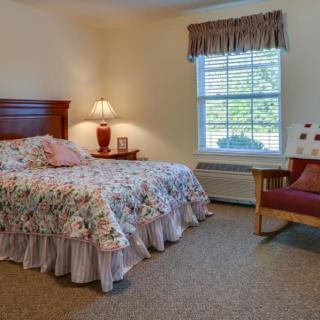 A cozy bedroom with a floral bedspread, lamp, and chair by the window.